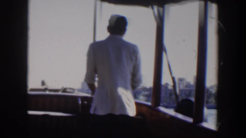 1951 Cuba home-movie frame showing a man in white viewed from behind at the helm of a boat near a bright waterfront.