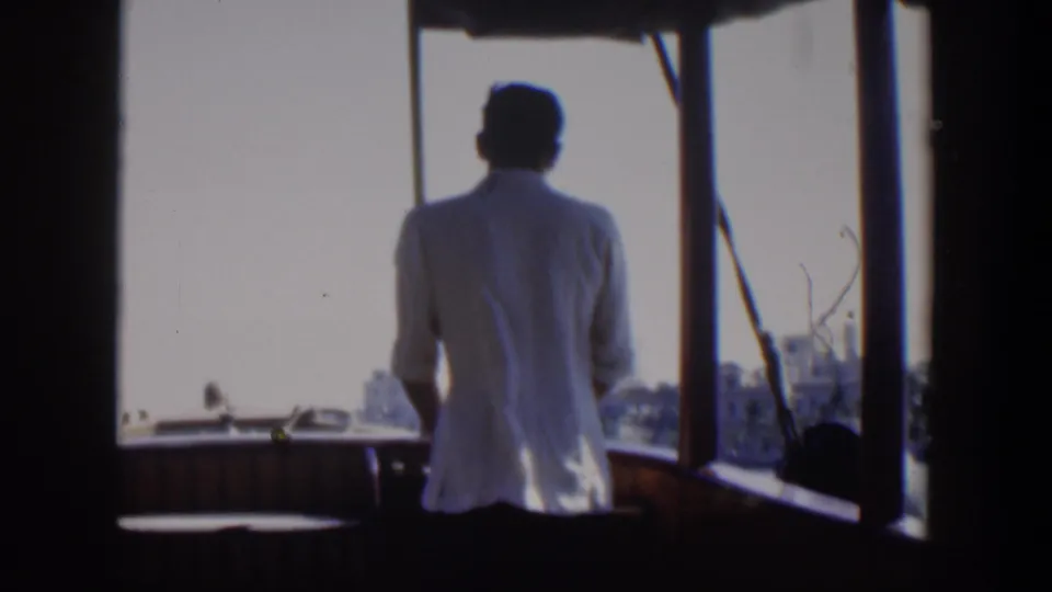 Rear view of the man at the boat helm, with Cuba waterfront buildings faintly visible beyond the sunlit water.