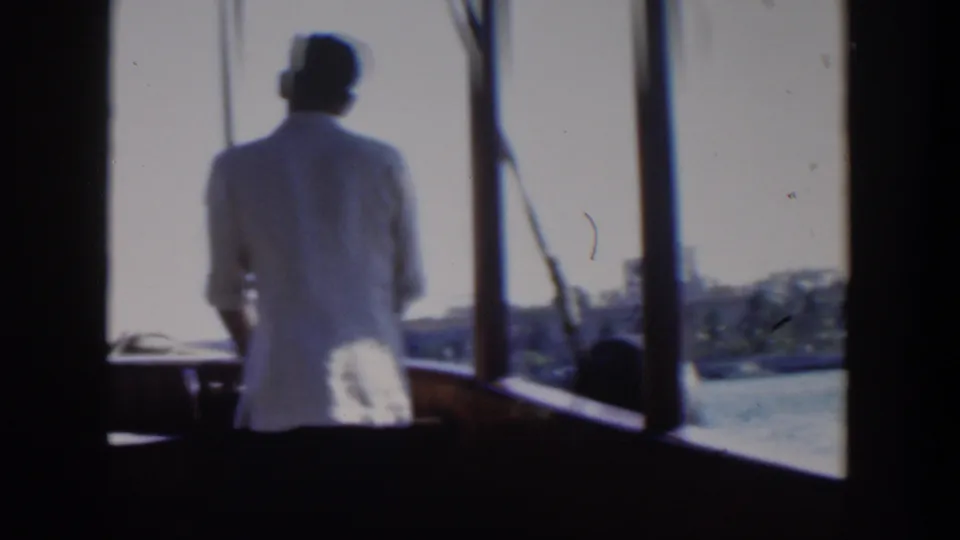 1951 Cuba boating frame showing the man at the helm, with bright water and a low urban shoreline visible ahead.