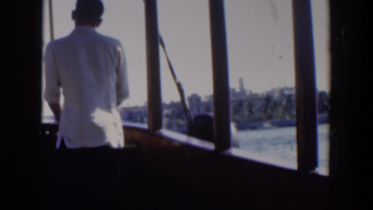 Man in a white shirt stands on a boat deck looking toward a distant shoreline.
