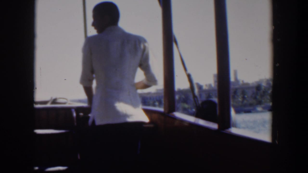 Man in a white shirt turns slightly on a boat as the coastline appears beyond him.
