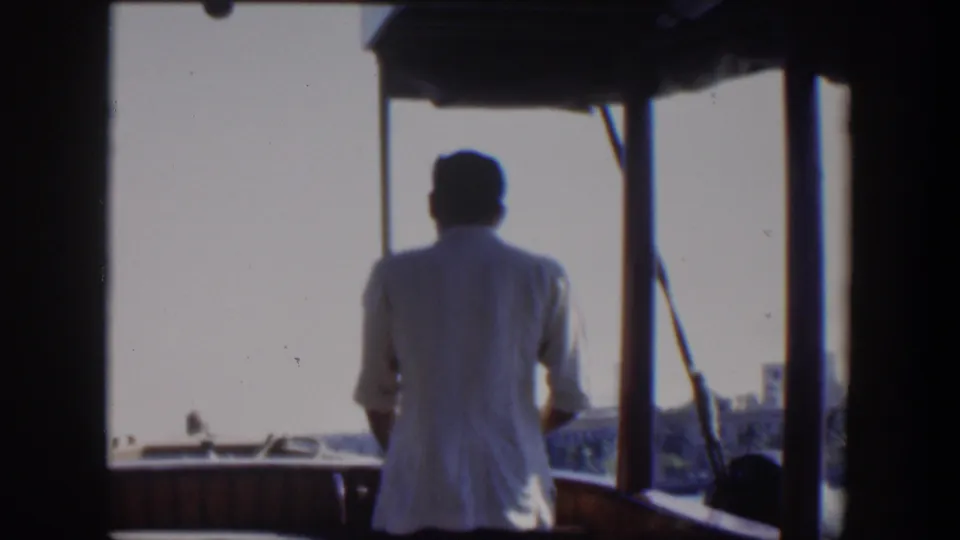 1951 Cuba 8mm frame showing a rear view of a man at the boat rail, framed by dark cabin posts and bright waterfront light.