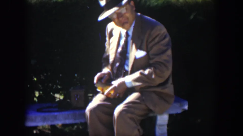 In a 1951 Cuba garden scene, a suited man in a fedora sits on a stone bench and looks down at a small yellow object.