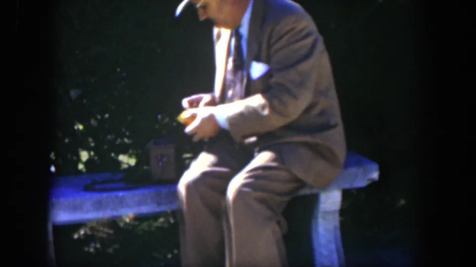 A 1951 Cuba 8mm frame shows the suited man in profile on a stone bench, still handling a small object in shaded garden light.