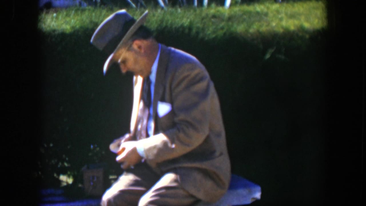 Man in fedora and light brown suit sits on a garden bench looking down at a small yellow package.