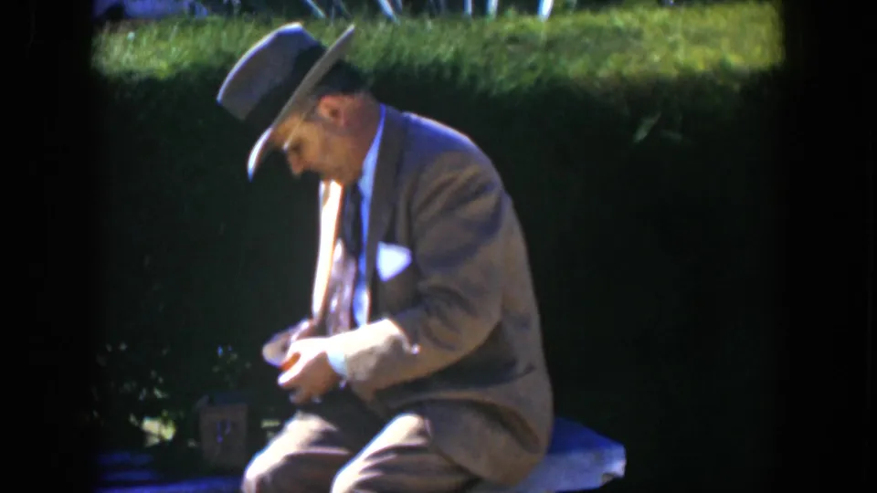 In 1951 Cuba home-movie footage, a suited man in a fedora sits outdoors in a garden and examines a small yellow package.