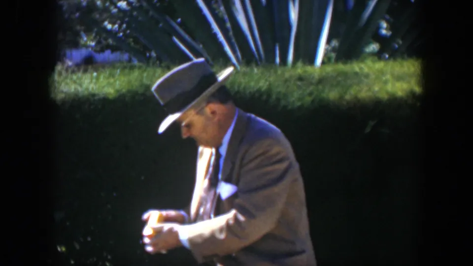 The man remains seated in the Cuban garden, holding the yellow package in both hands beneath bright sunlight.