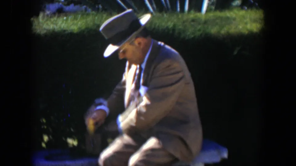 In the garden setting, the suited man leans forward as he lowers or adjusts the small yellow package near the bench.