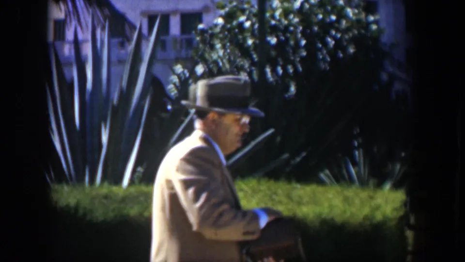 The 1951 Cuba home-movie frame shows the suited man turned sideways on the bench in a sunlit tropical garden.