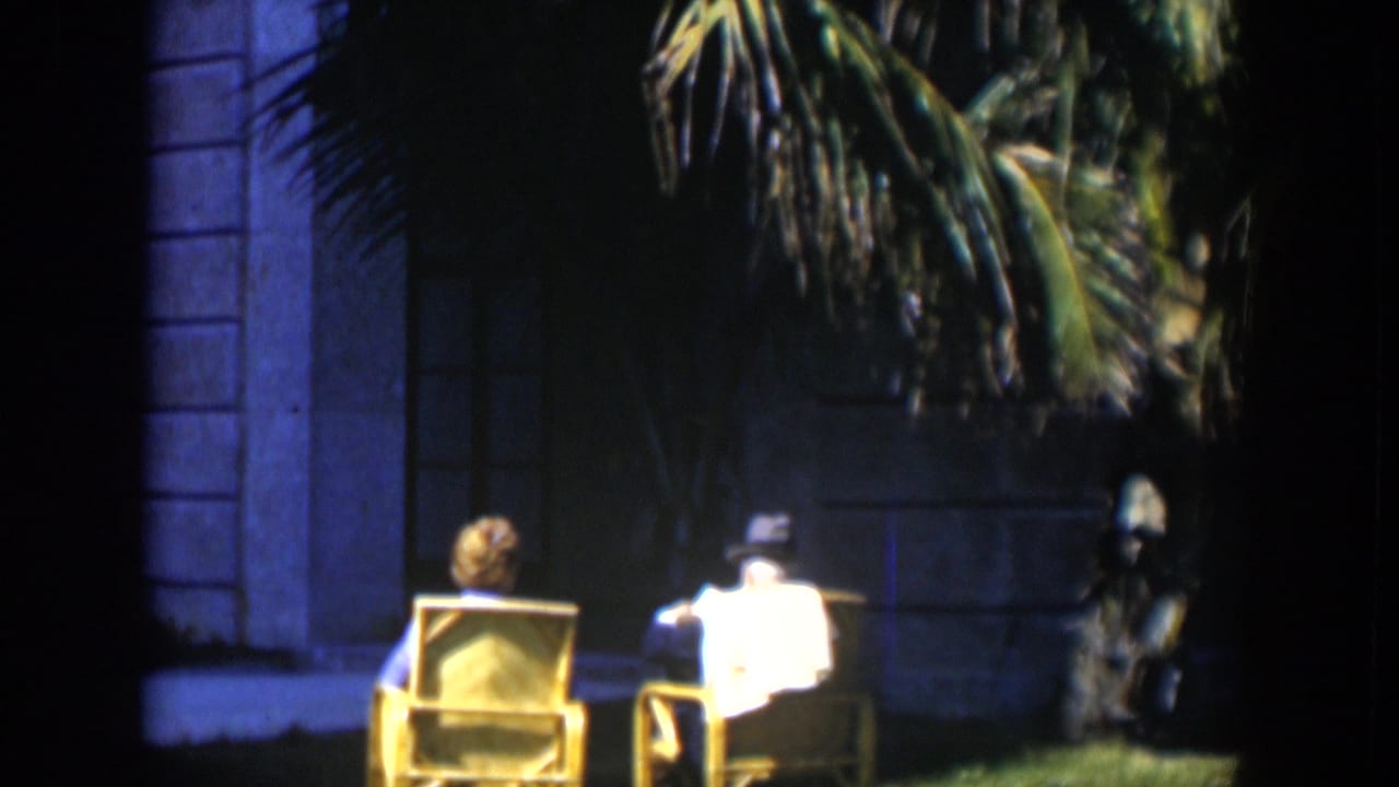 In 1951 Cuba, two adults sit in wicker lawn chairs outside a stately building, partly shaded by palm fronds.