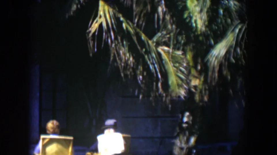 A 1951 Cuba home-movie frame shows two adults seated outdoors near a palm and the facade of a large residence.