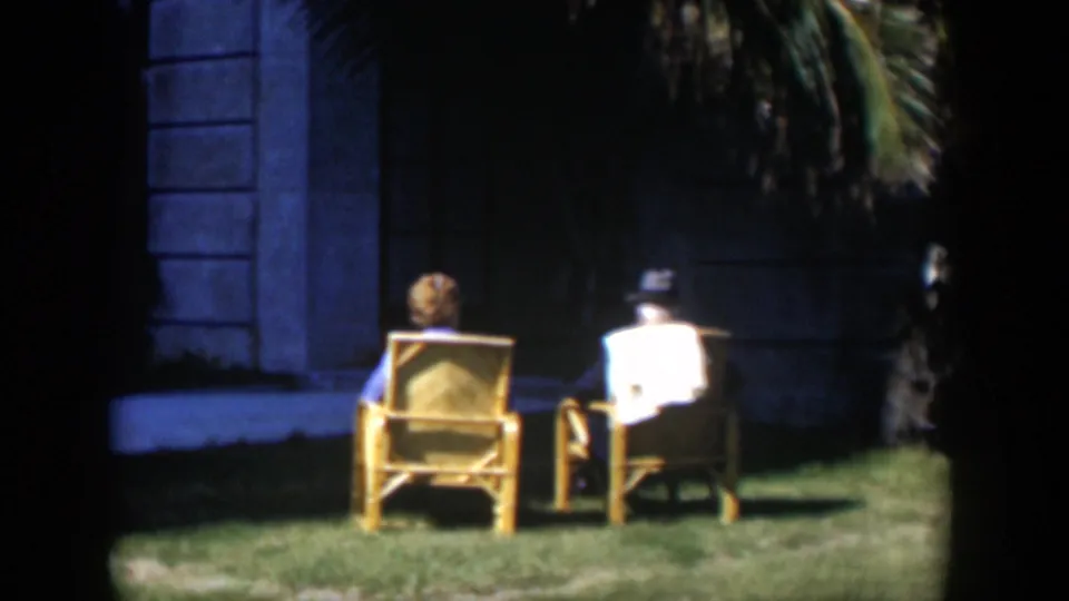 In the Cuba 1951 clip, the couple remains seated in wicker chairs on grass, facing the shaded mansion wall and palm foliage.