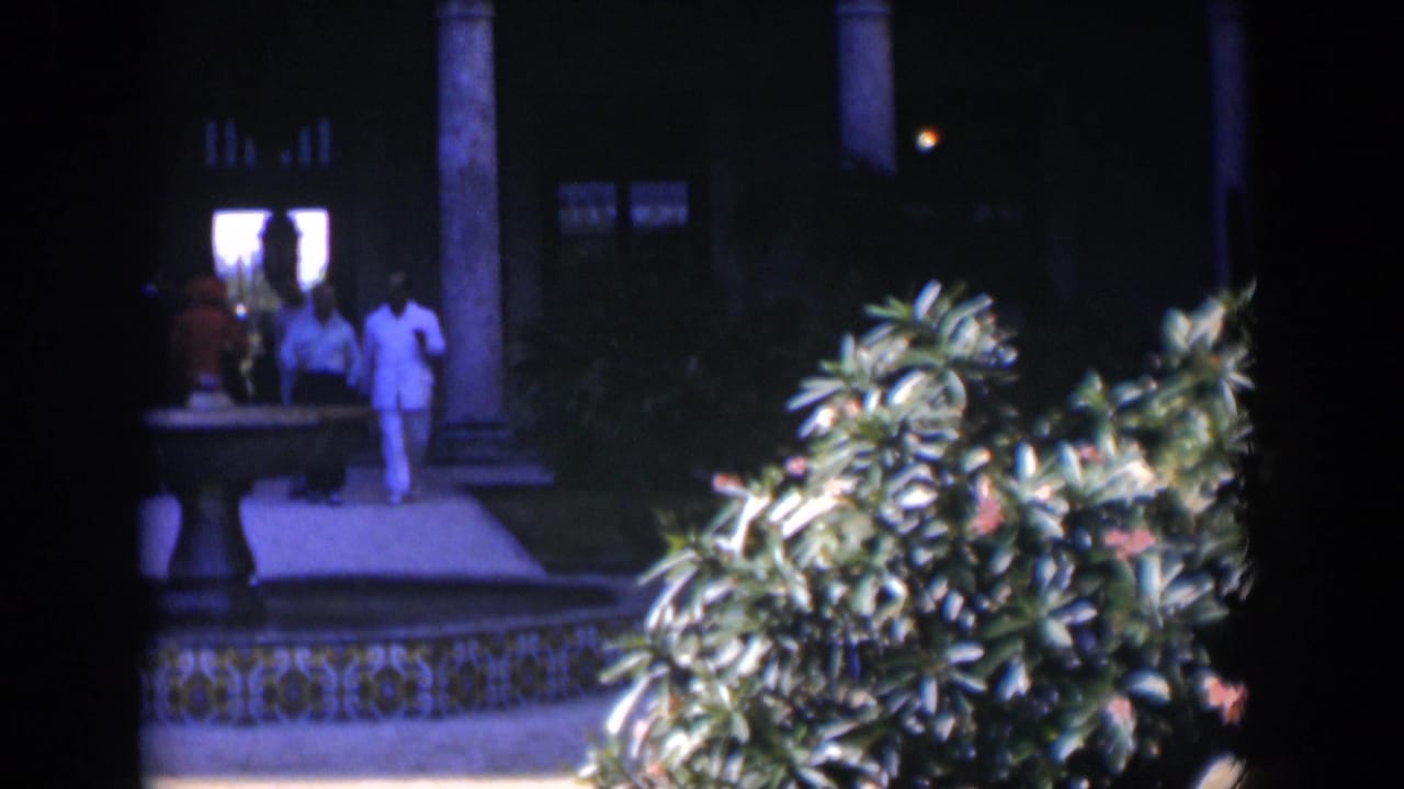 Men in light clothing walk near a tiled fountain in a shaded courtyard.