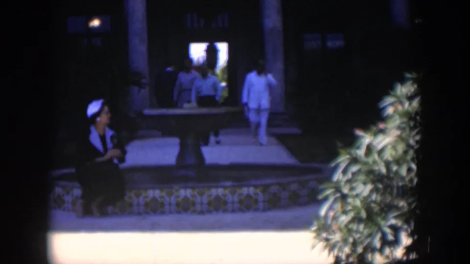 1951 Cuba 8mm frame with a seated woman in a white headscarf near the tiled fountain while men pass in the background.