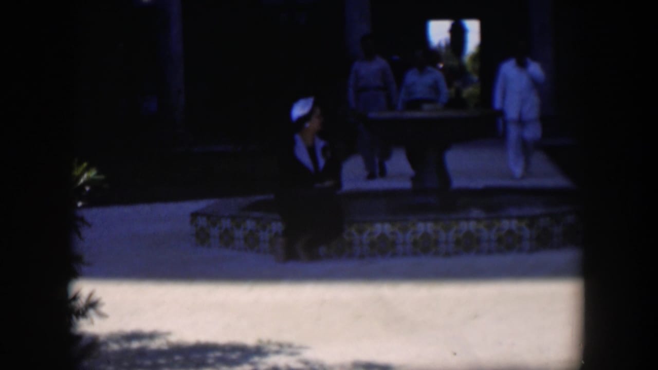 People walk near a shaded courtyard while a seated woman is visible at the left edge.