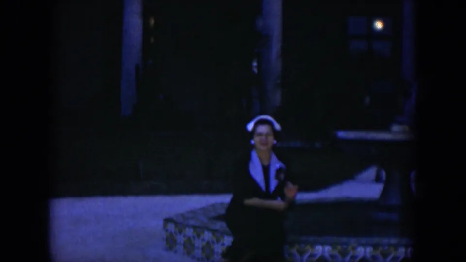1951 Cuba 8mm frame showing a woman in a dark dress and white headscarf seated on a patterned bench in a shaded courtyard.