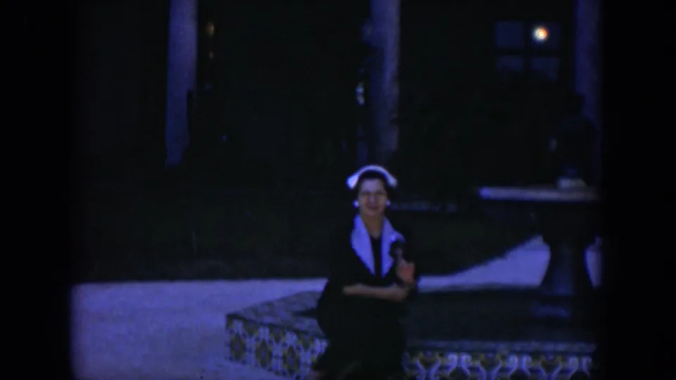 1951 Cuba 8mm frame of a seated woman wearing a white headscarf on a tiled bench, framed by shaded columns and dim courtyard space.
