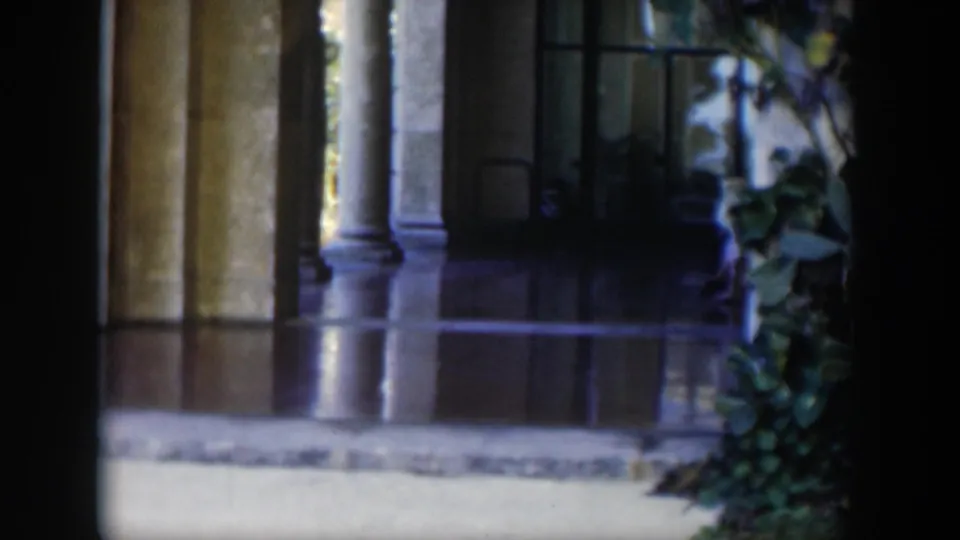 A soft 8mm view of a Cuban building exterior shows columns, dark window openings, reflective flooring and nearby foliage.