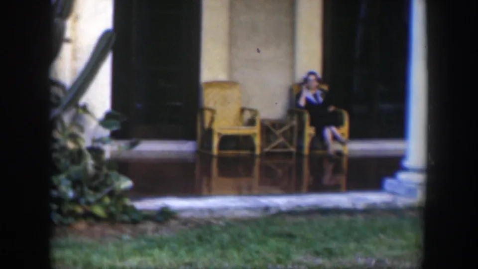 1951 Cuba frame showing a woman seated on a tiled porch near wicker chairs, with tropical plants and a doorway behind her.