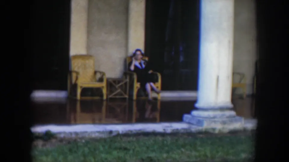 1951 Cuba 8mm frame with a seated woman on a tiled veranda near a white column, wicker furniture, and shaded doorways.