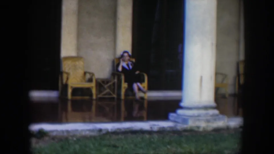 In 1951 Cuba footage, a woman sits on a tiled porch near wicker furniture and large columns.