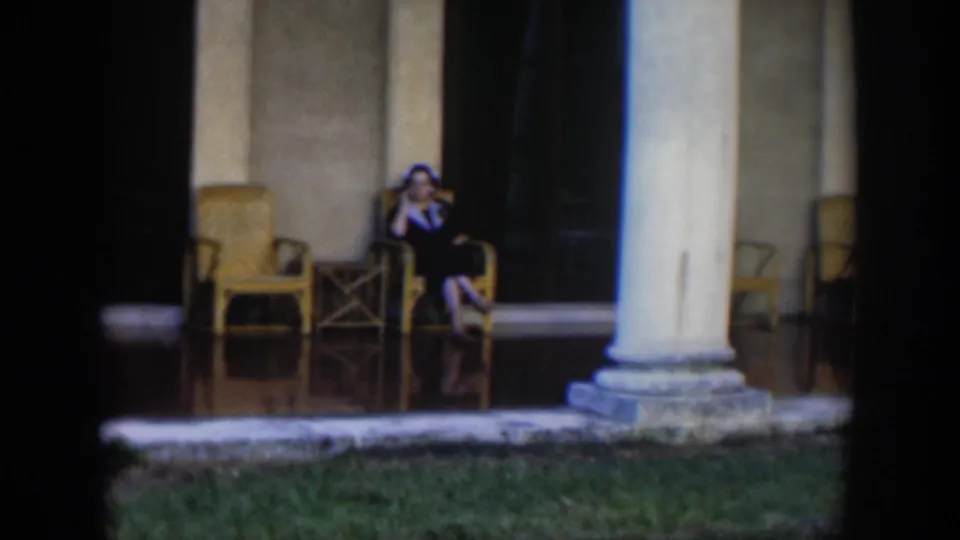 The woman remains seated on the porch, framed by pale columns, wicker chairs, and a dark doorway.