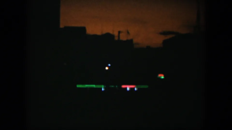 A dark night view shows silhouetted rooftops beneath an orange sky with small red, green, and blue lights.