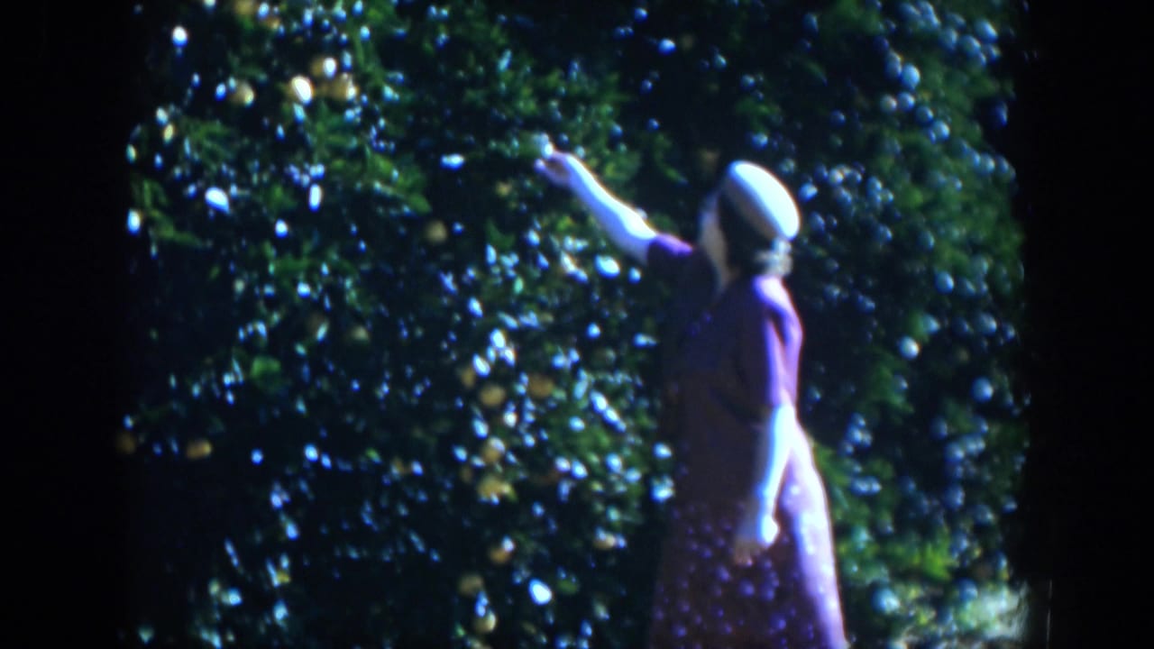 Woman stands beside dense foliage with her arm raised toward fruit.
