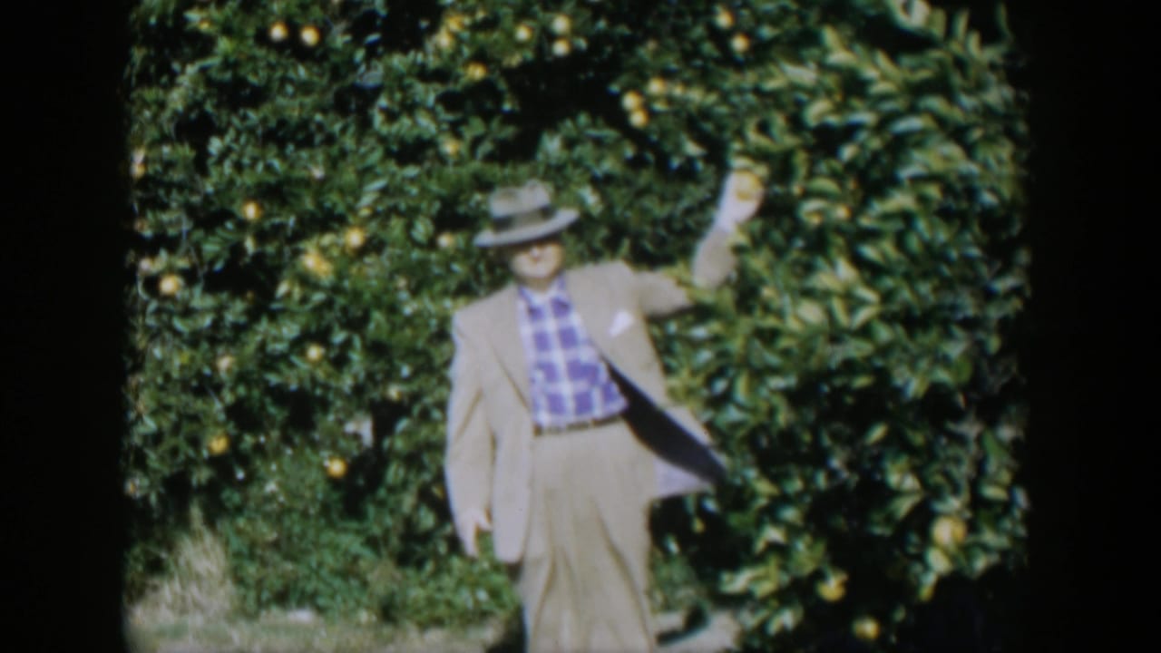 Man in a light suit and fedora stands before citrus trees with one arm raised.
