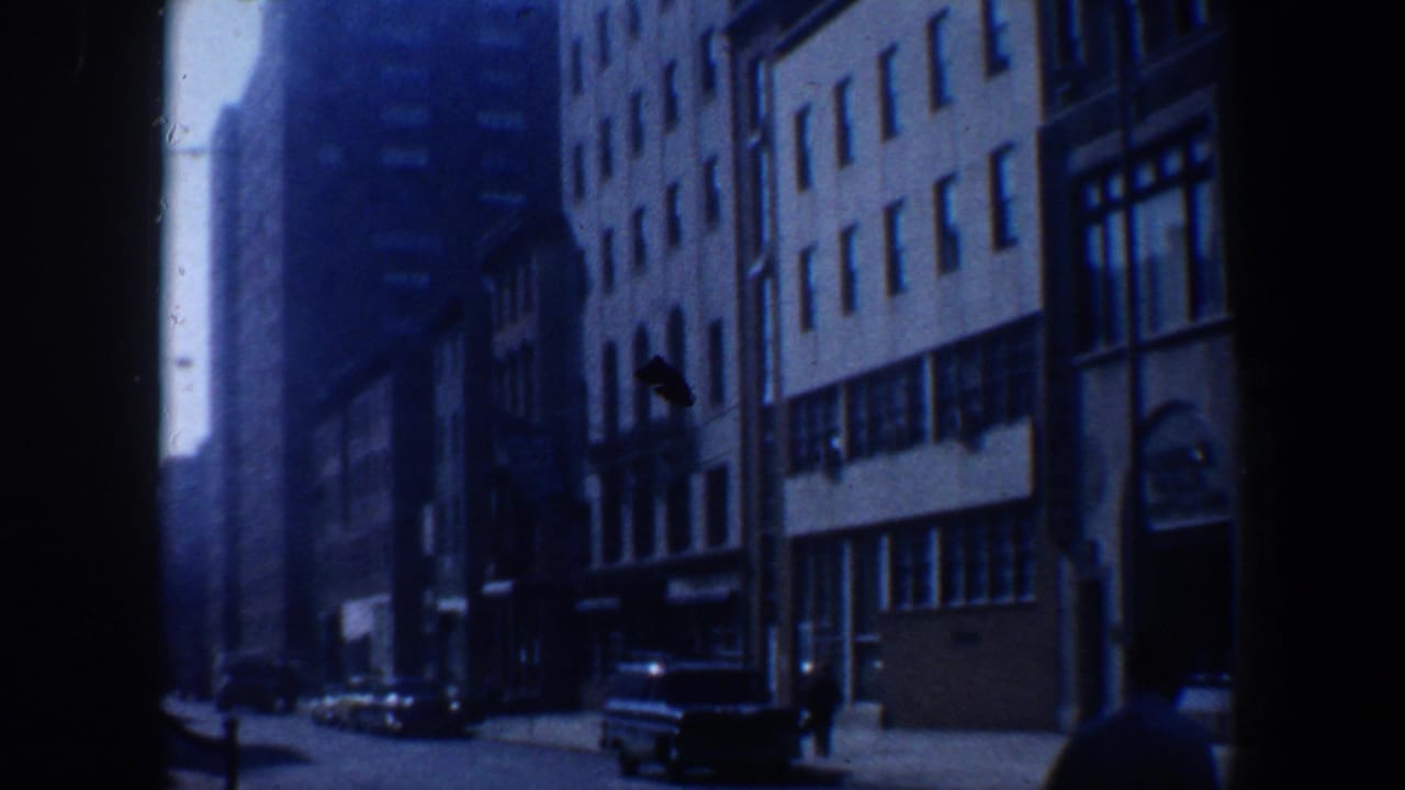 Blue-tinted street view with multi-story buildings, parked cars, and a distant pedestrian.