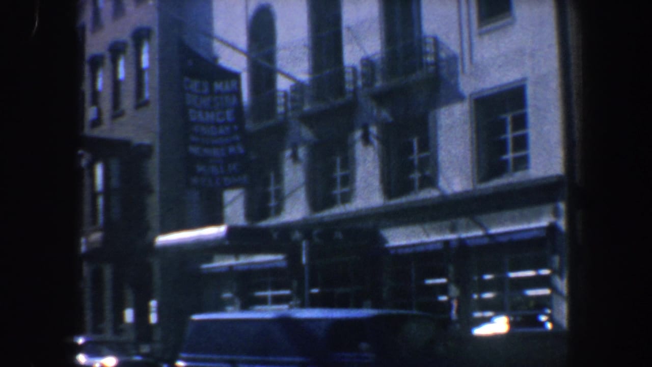 Street-level view of an urban building facade with awnings, fire escapes, a hanging sign, and a car at the curb.