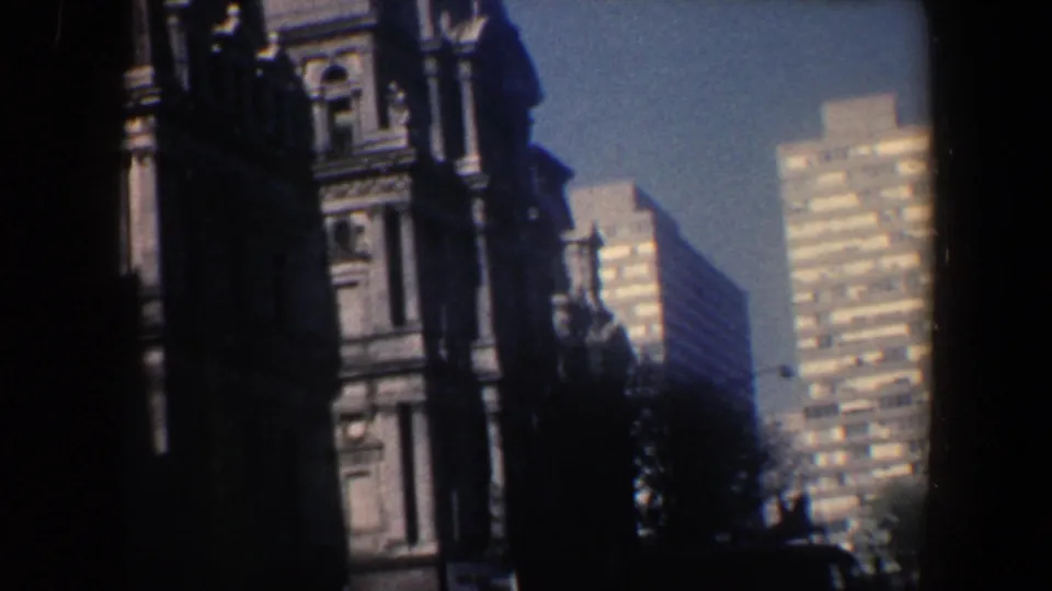 1962 Philadelphia 8mm frame showing a dark ornate building edge, tree shadows, and a tall sunlit high-rise in the background.