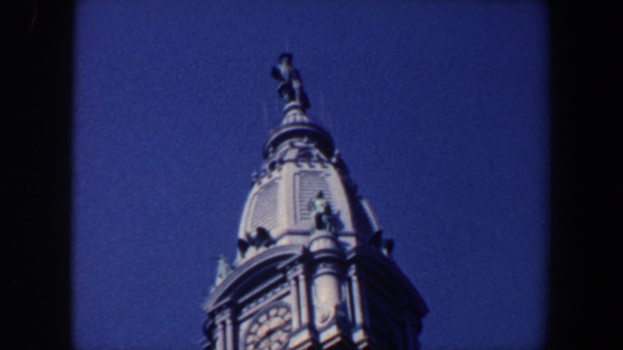 1962 Philadelphia 8mm frame showing the upper portion of an ornate clock tower with a rooftop statue against blue sky.