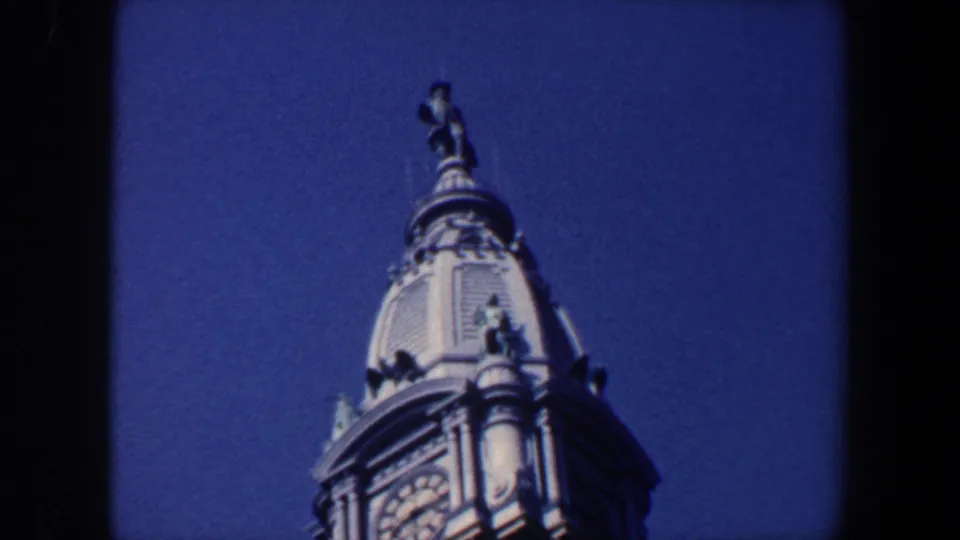 1962 Philadelphia 8mm frame showing the upper portion of an ornate clock tower with a rooftop statue against blue sky.