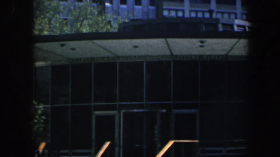 Philadelphia 1962 8mm frame showing a modernist public hall entrance with curved glass, steps, railings, trees, and heavy shadow framing.