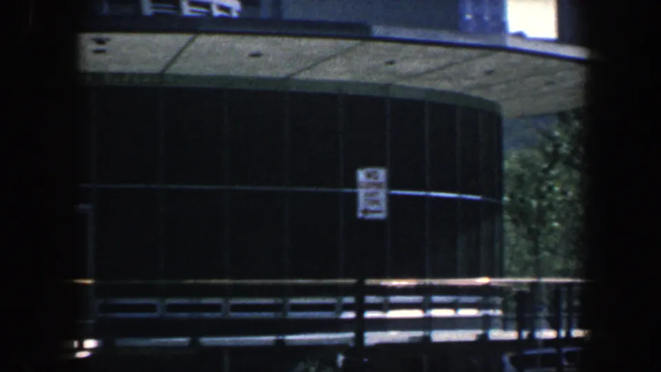 Philadelphia 1962 8mm frame showing the rounded glass side of the hall, a street sign, sidewalk, trees, and railings under strong shadow framing.