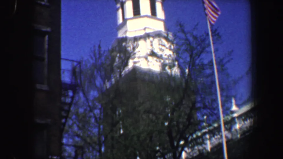1962 Philadelphia frame showing the upper tower of a historic brick building with white trim, trees, a flagpole, and clear blue sky.