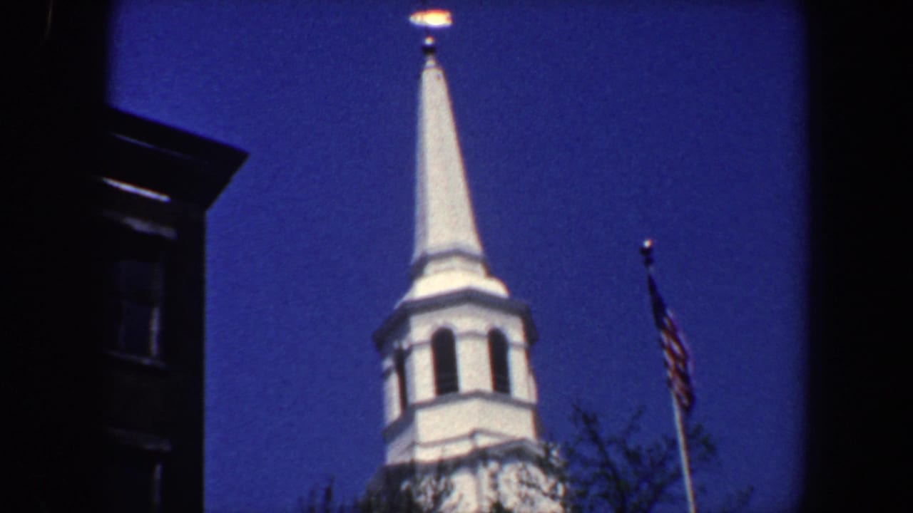 Church spire and American flag against a deep blue sky with dark building edges.