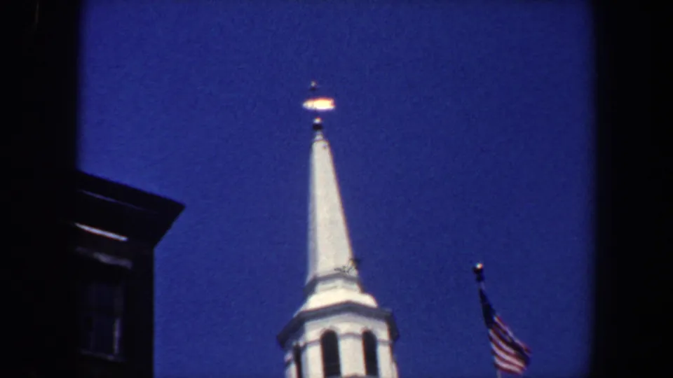 1962 Philadelphia home-movie frame emphasizing the pointed white spire, nearby American flag, and dark urban building silhouette.