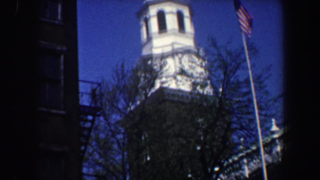 White church steeple above trees with an American flag beside dark brick buildings.