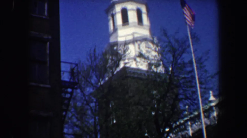 1962 Philadelphia home-movie frame showing a white church steeple, trees, dark brick buildings, and an American flag on a pole.