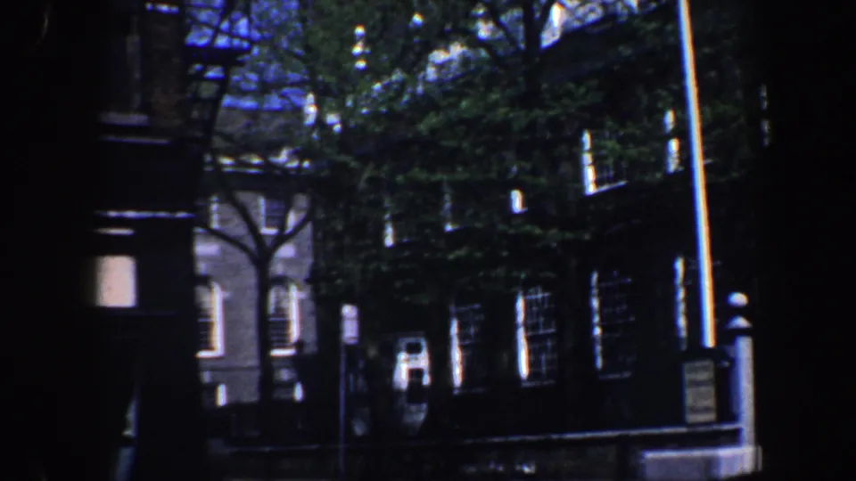 1962 Philadelphia exterior showing brick windows, leafy trees, a flagpole, and a small sidewalk sign in a handheld 8mm view.