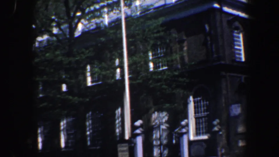 Philadelphia home-movie frame with a flagpole centered before brick buildings, trees, windows, and a projecting roofline.