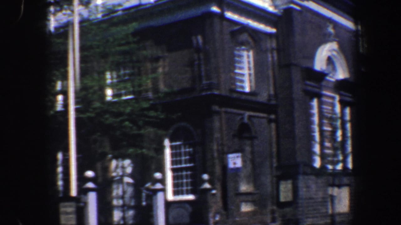 Brick building corner with arched windows, white trim, flagpole, and trees in partial shade.