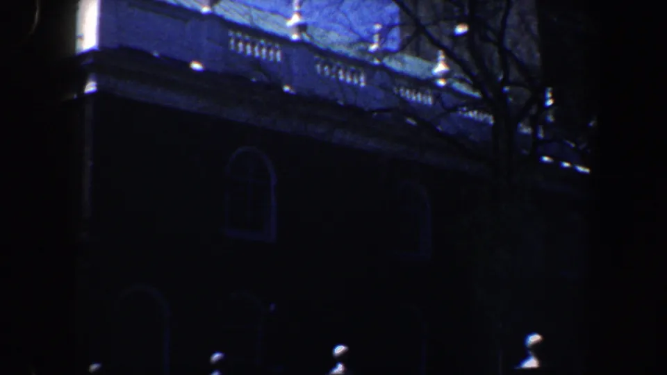 1962 Philadelphia frame showing the shadowed side facade with rooftop balustrade, arched windows, bare branches, and small bright forms near the base.