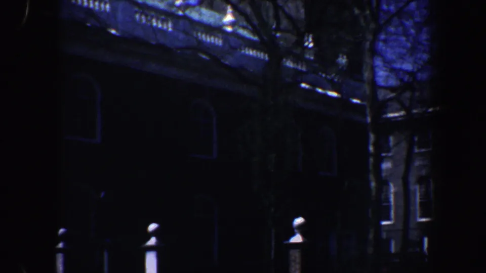 1962 Philadelphia 8mm frame showing a dim older brick building partly obscured by bare trees and deep blue shadow.