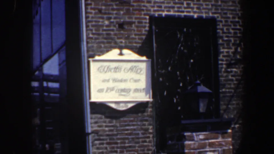 1962 Philadelphia frame showing a readable painted sign on a brick wall, with a dark window and exterior stair nearby.