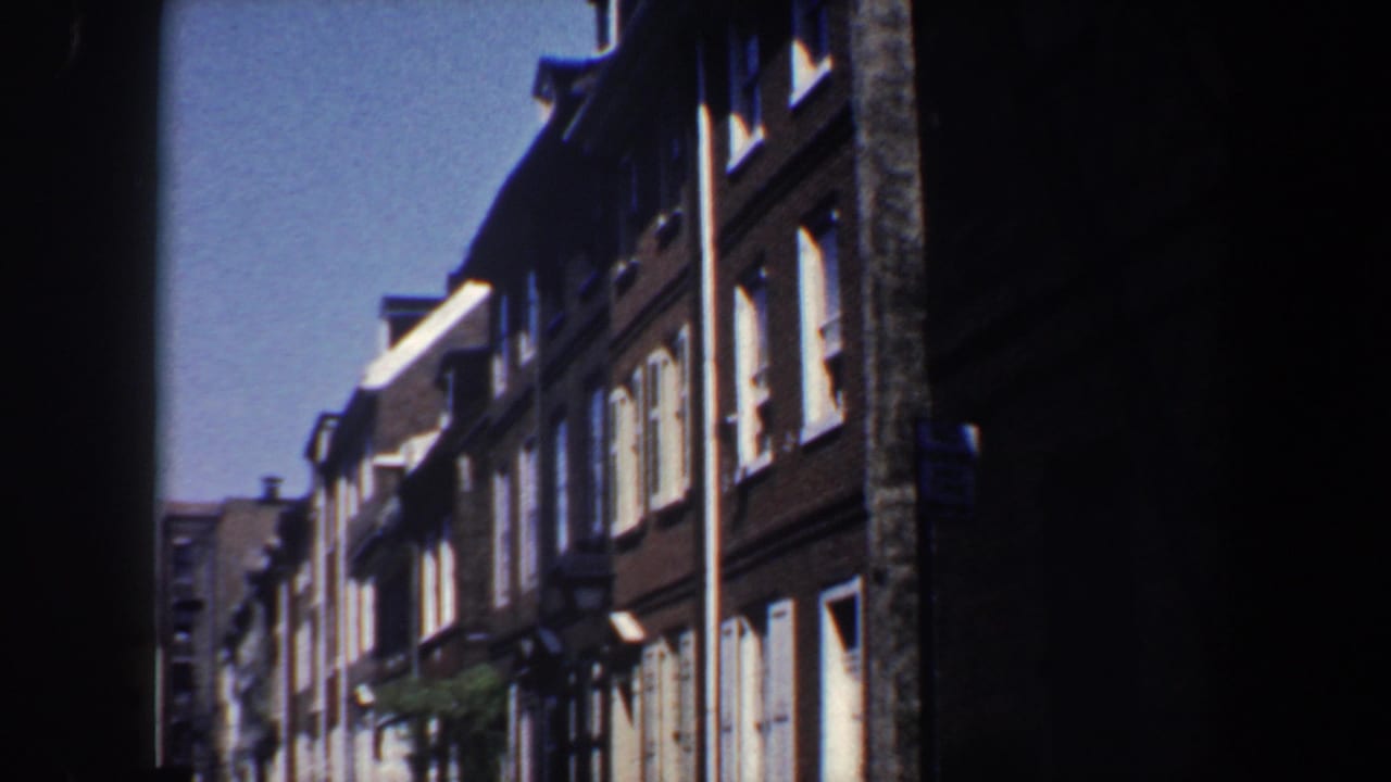 1962 Philadelphia home-movie frame looking down a narrow residential street lined with brick row houses, white shutters, and deep foreground shadows.