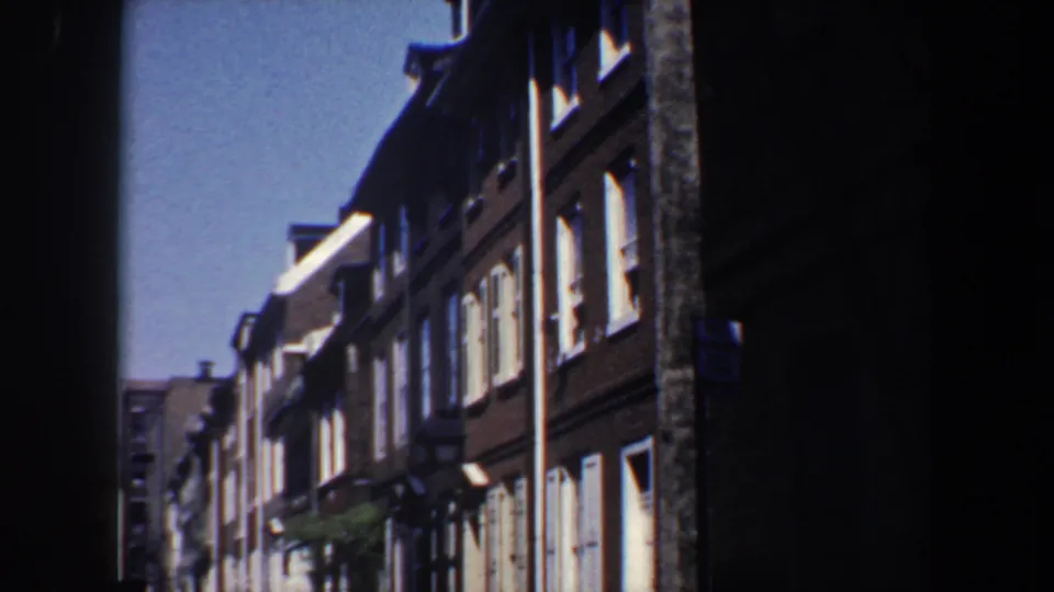 1962 Philadelphia home-movie frame looking down a narrow residential street lined with brick row houses, white shutters, and deep foreground shadows.