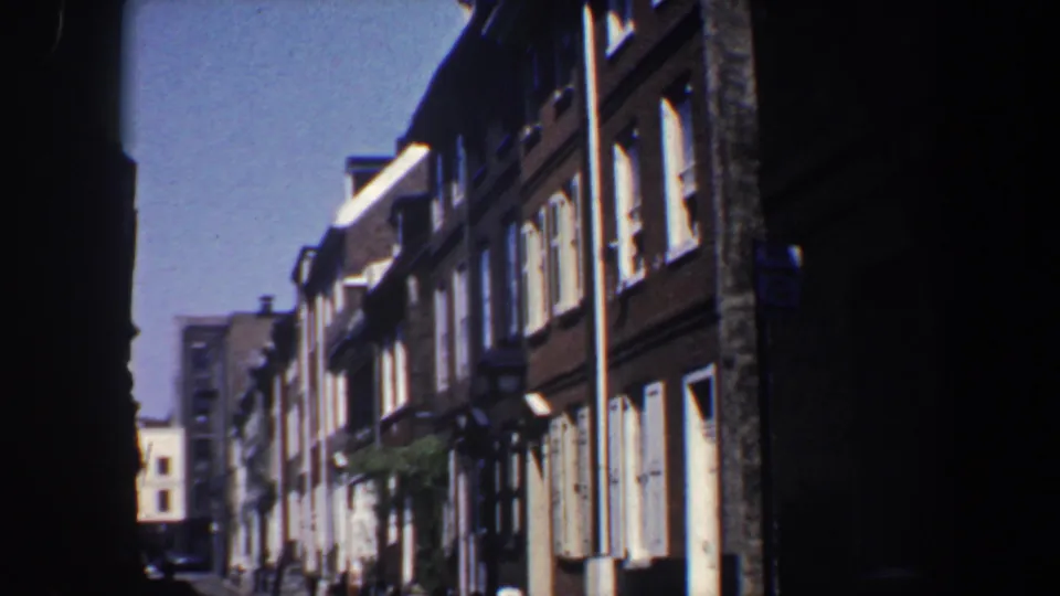 1962 Philadelphia frame showing a low-angle view of brick row houses, pale shutters, street lamps, and a distant building at the end of the block.